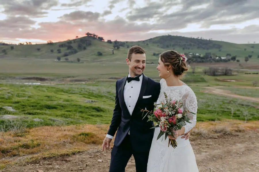 Bride and groom smiling on a grassy hillside at dusk.