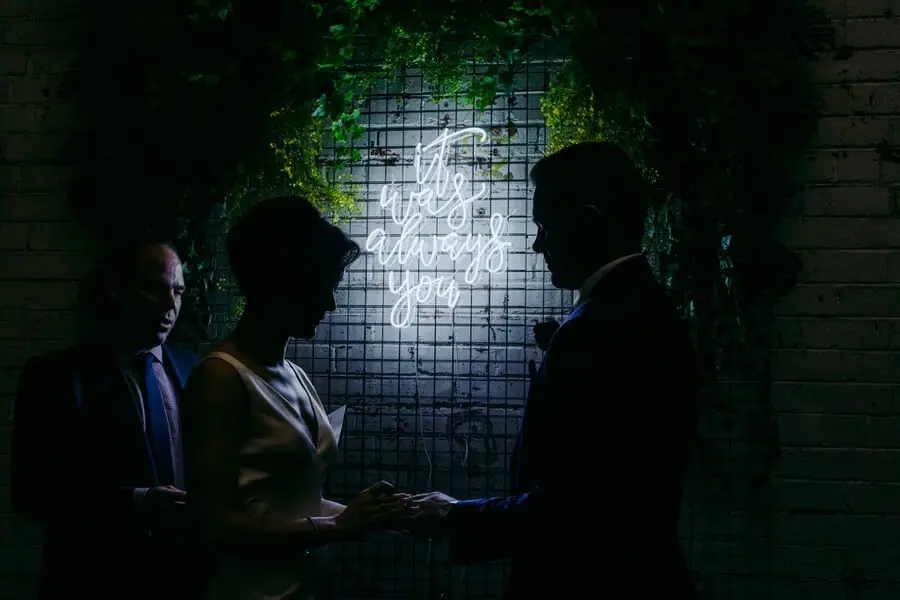 Couple holding hands in silhouette beneath a glowing wall sign.
