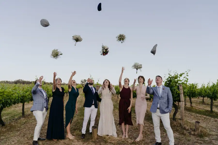Wedding party throwing bouquets and hats above vineyard rows.