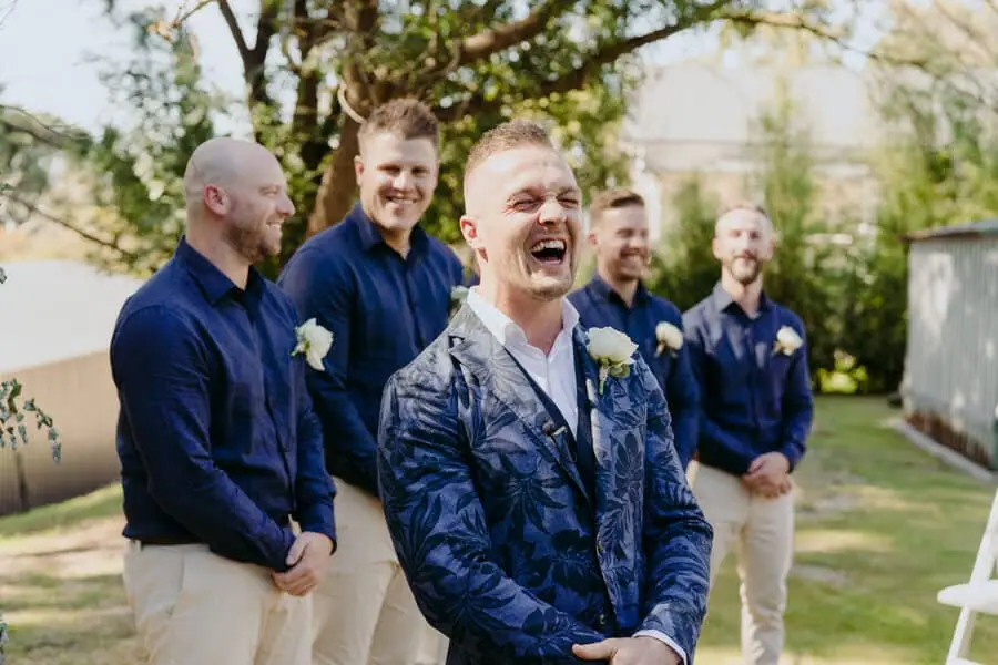 Groom laughing with companions beneath garden trees.