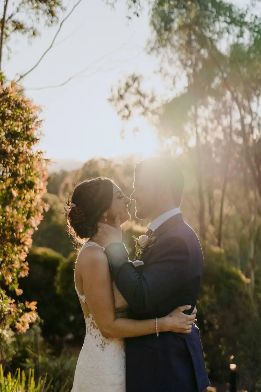 Bride and groom embracing in warm sunlight beneath leafy trees.