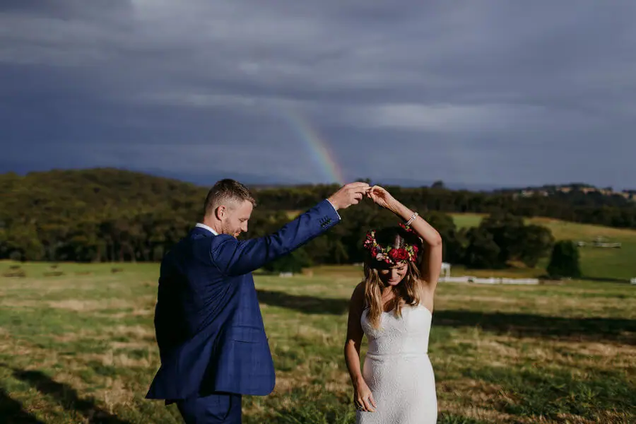 Groom turning the bride beneath a cloudy sky and a rainbow.
