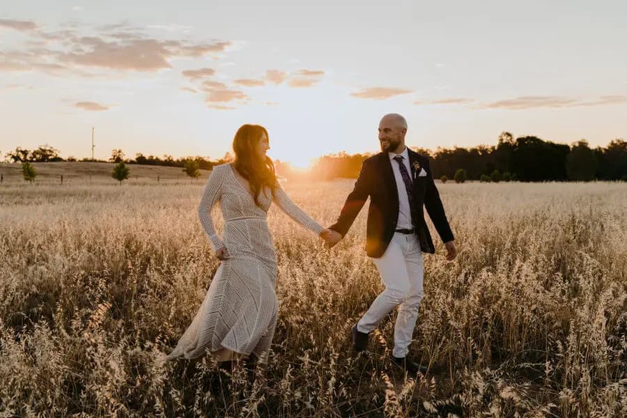 Bride and groom walking hand in hand through tall grass at sunset.