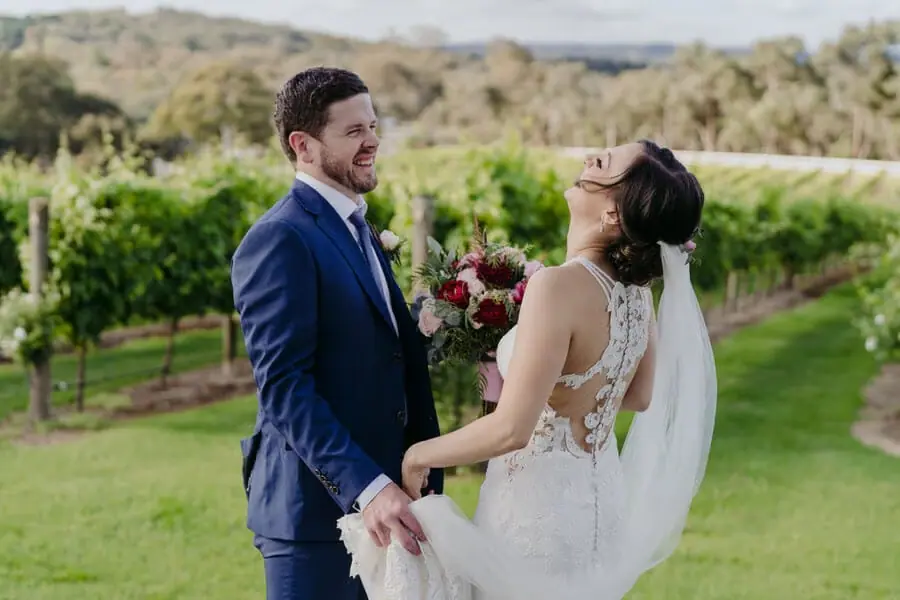Bride laughing while holding hands with the groom beside grapevines.