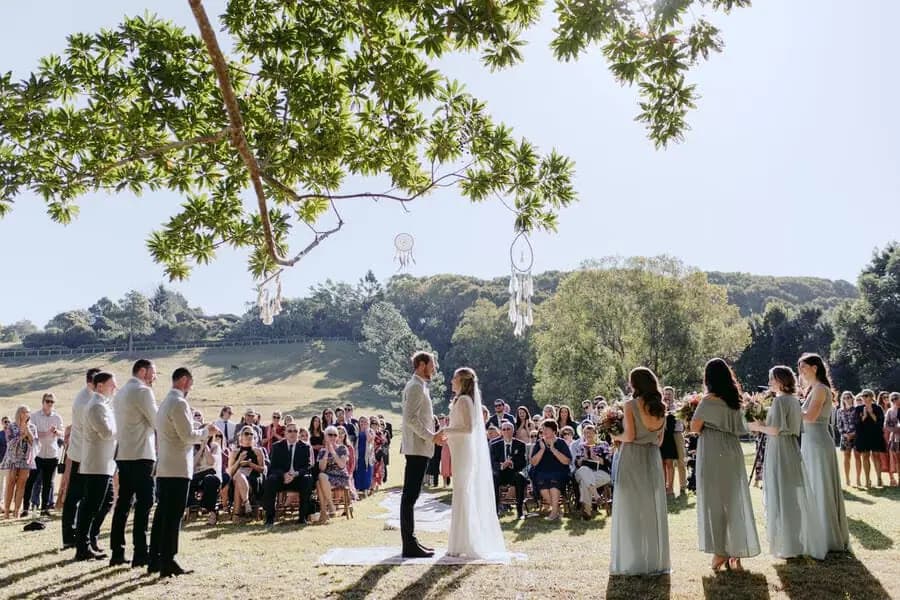 Wedding party and guests gathered at an outdoor ceremony beneath a tree.