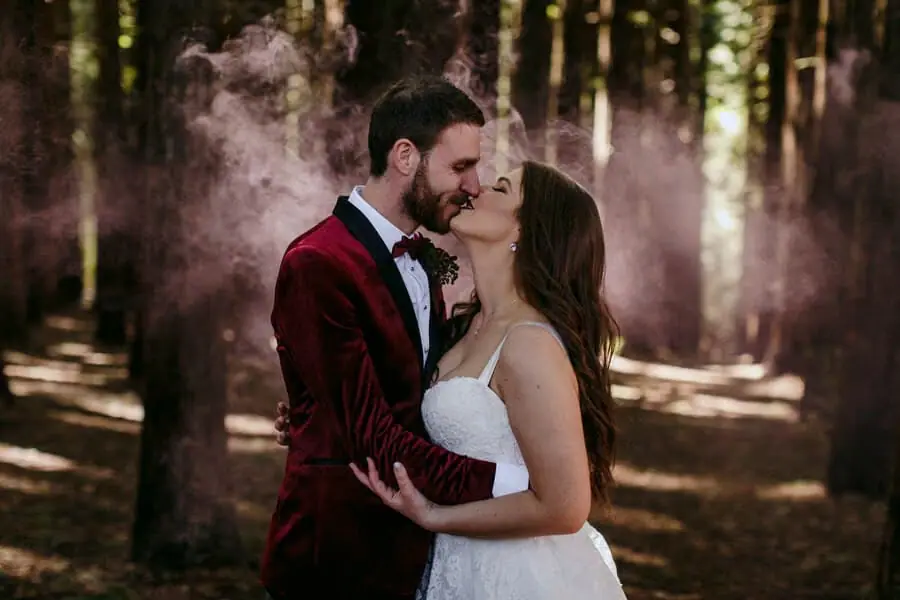Couple kissing among forest trees with pink haze behind them.
