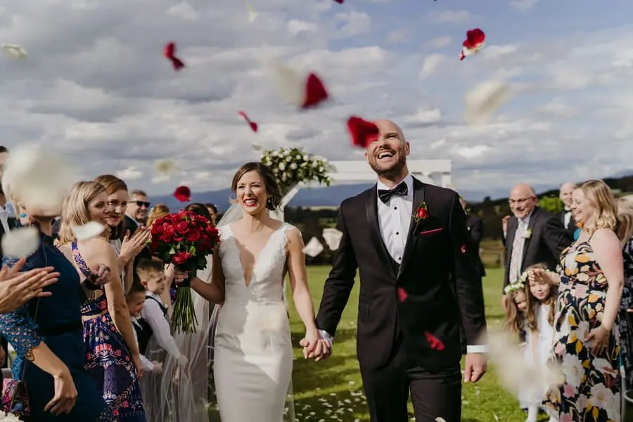 Couple smiling as guests throw red petals over them.