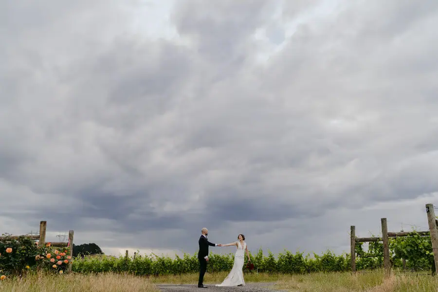 Couple holding hands beneath a wide cloudy sky beside grapevines.