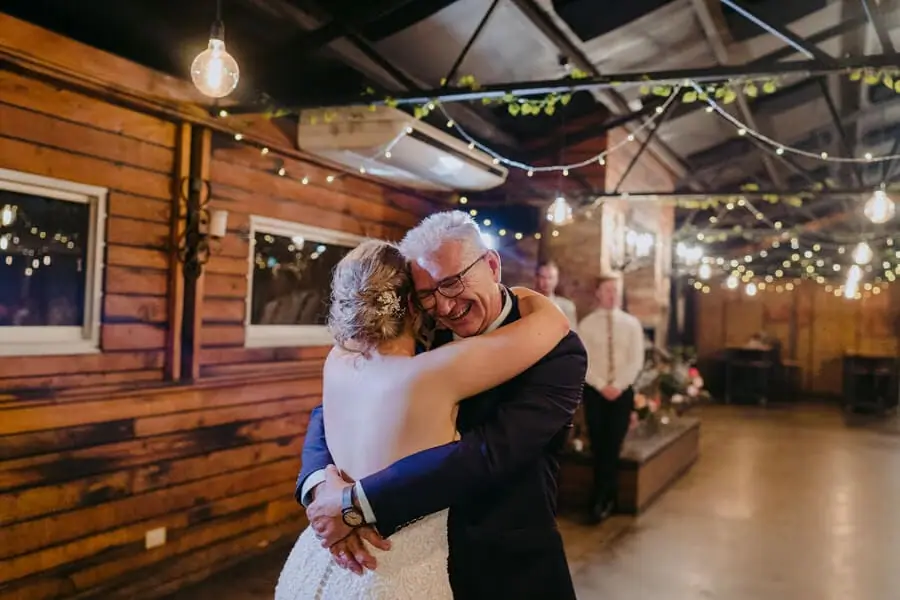 Bride hugging a man in glasses beneath reception string lights.