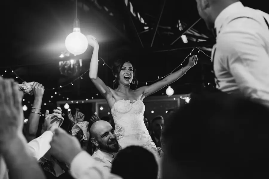 Black-and-white photograph of a bride raising both arms on a crowded dance floor.