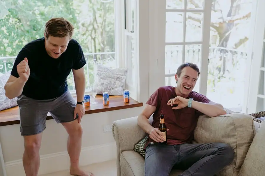 Two men laughing together beside a sofa and bright windows