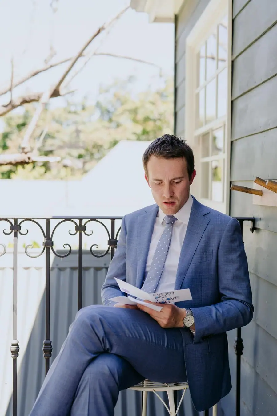 Groom reading a notebook while seated on a veranda in a blue suit
