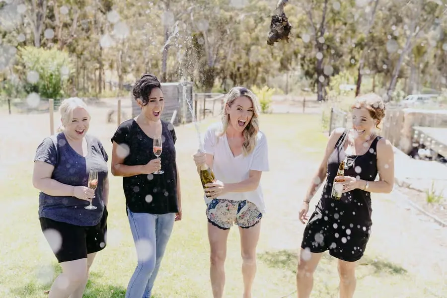 Four women laughing together as a bottle sprays sparkling wine on a lawn