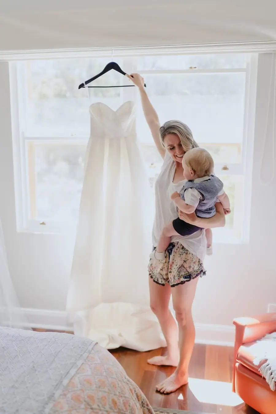 Bride holding a baby while lifting her wedding dress on a hanger beside a window