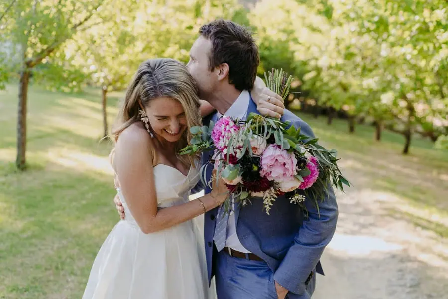 Groom kissing the laughing bride's forehead beneath garden trees