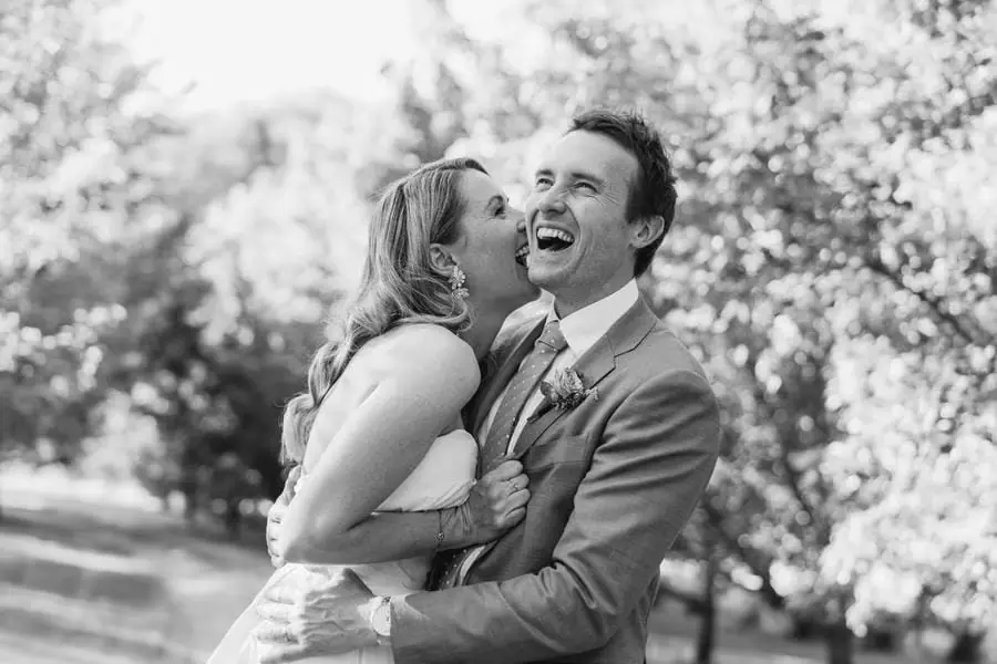 Black and white photograph of the bride kissing the laughing groom's cheek