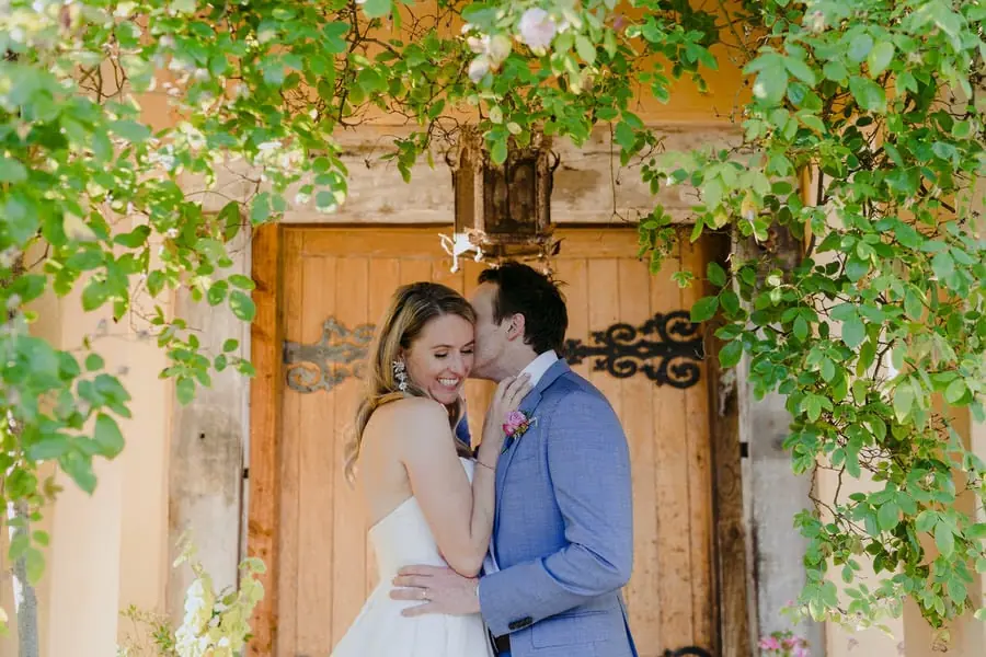 Groom kissing the bride's temple beside a wooden door covered with vines