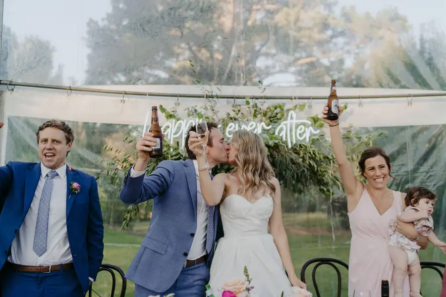 Bride kissing the groom as the wedding party raise their drinks