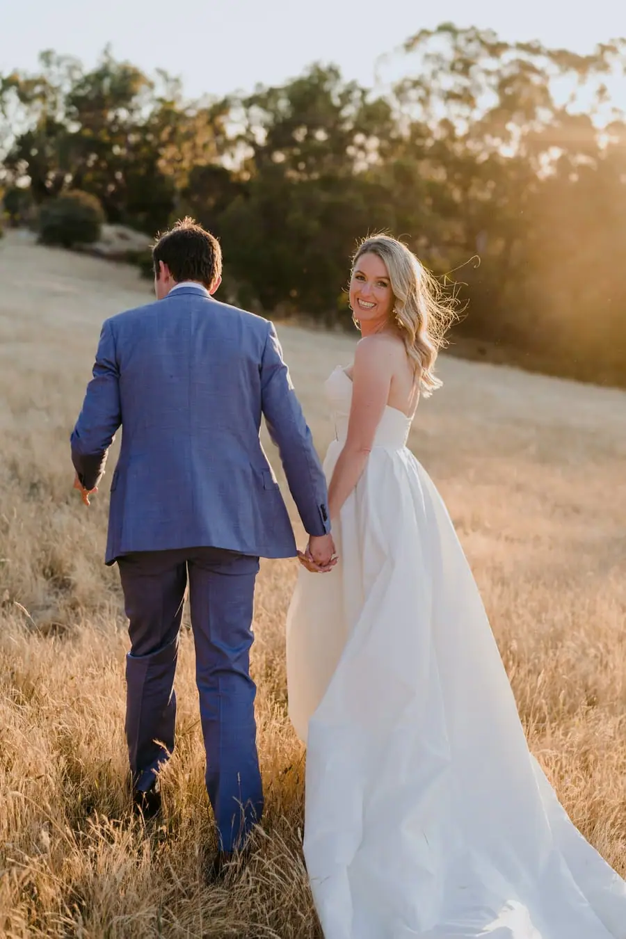 Bride and groom holding hands while walking through golden grass at sunset