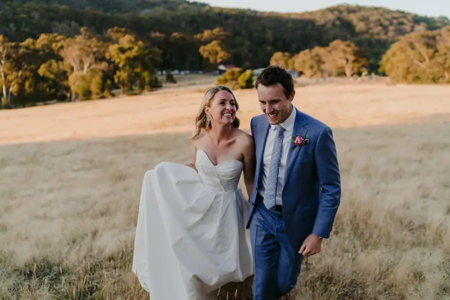 Bride and groom laughing while walking through a sunlit field