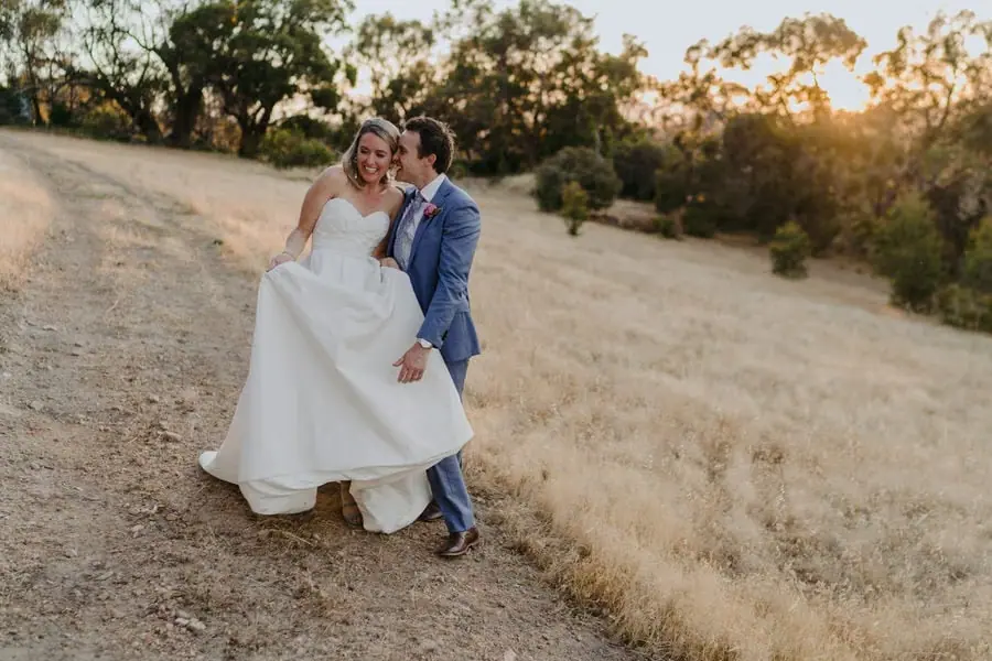 Bride and groom walking closely together through dry grass at sunset