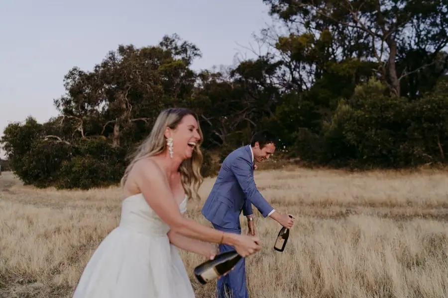 Bride and groom laughing together while holding bottles in a field