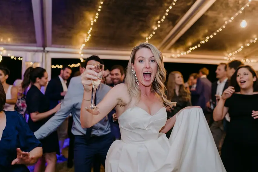 Bride cheering toward the camera while holding a drink and lifting her skirt