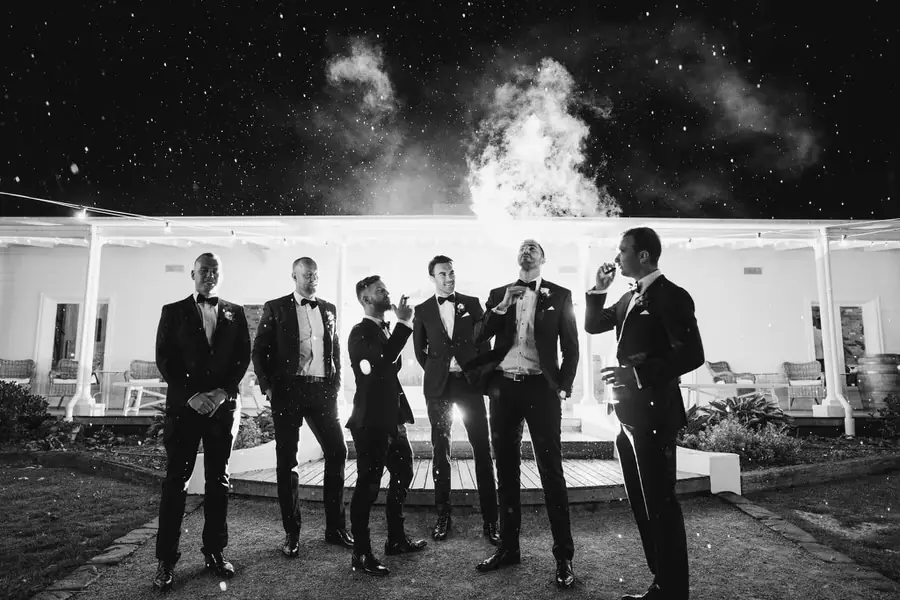 Black and white photograph of men in suits standing beneath nighttime lights and airborne spray.