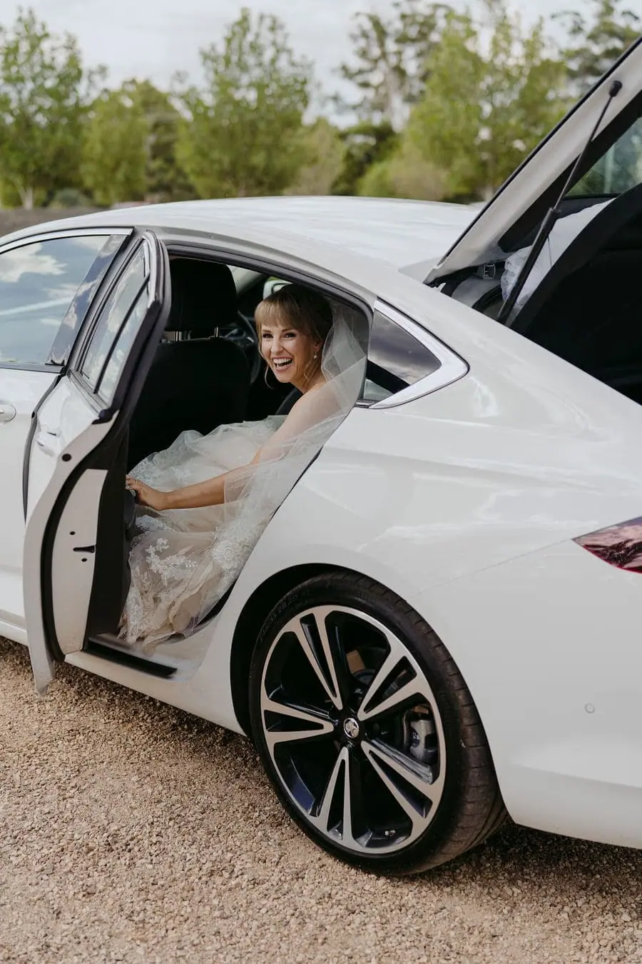 Bride sits in the open rear door of a white car.