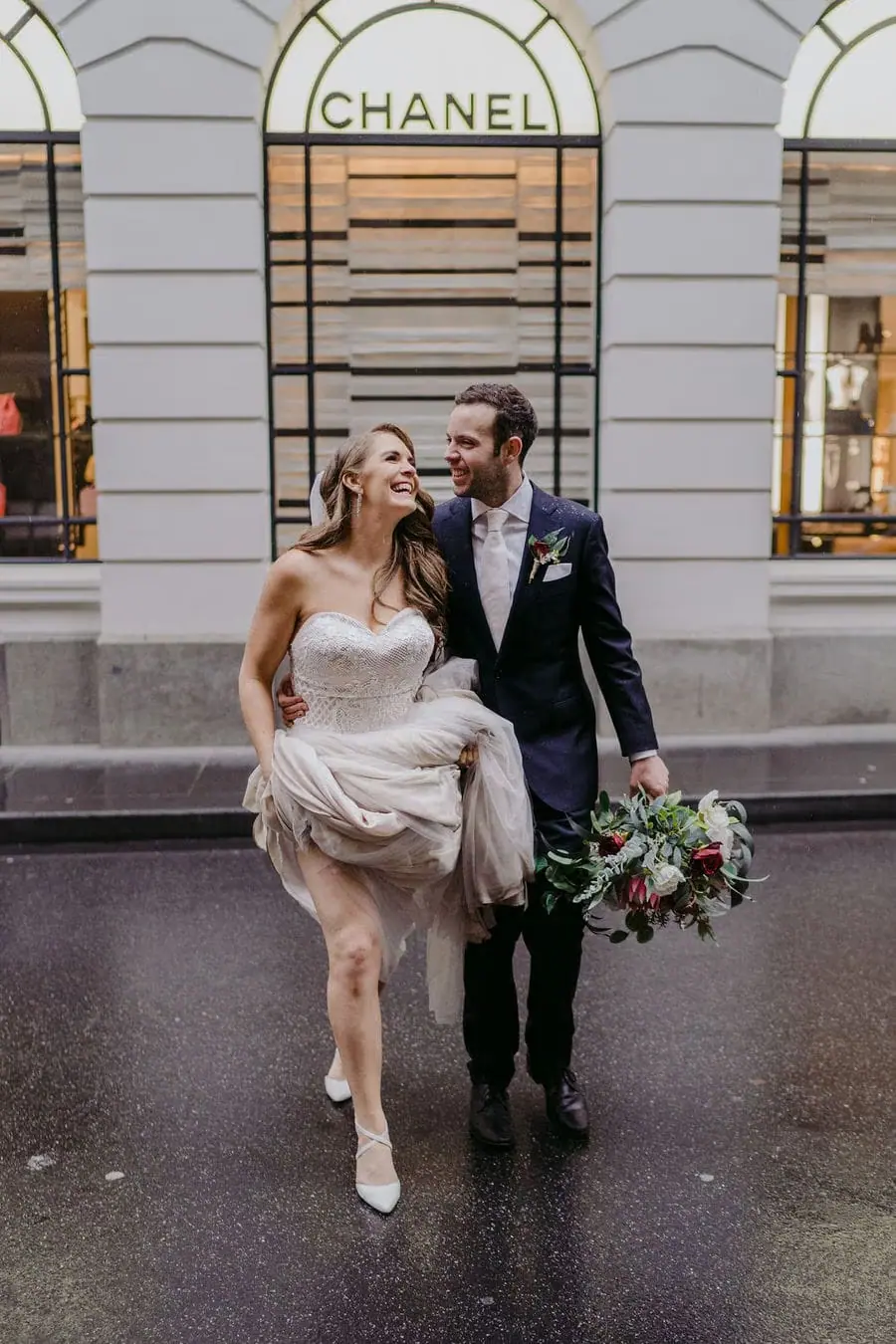 Couple walks outside a shopfront with the bride lifting her skirt and holding flowers.