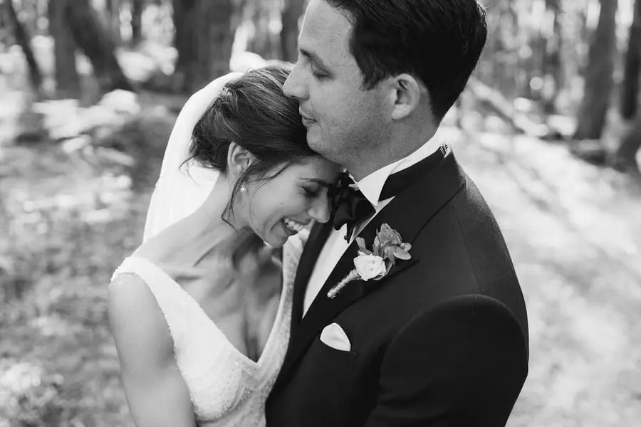 Black and white photograph of the groom kissing the bride's forehead among trees.