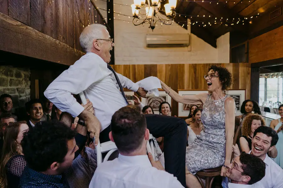 Guests lift an older man in a chair while a woman celebrates beside him.