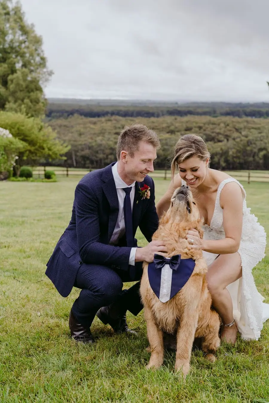 Couple crouches beside a dog on a lawn.
