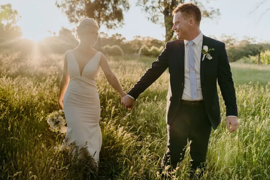 Couple walks hand in hand through sunlit long grass.