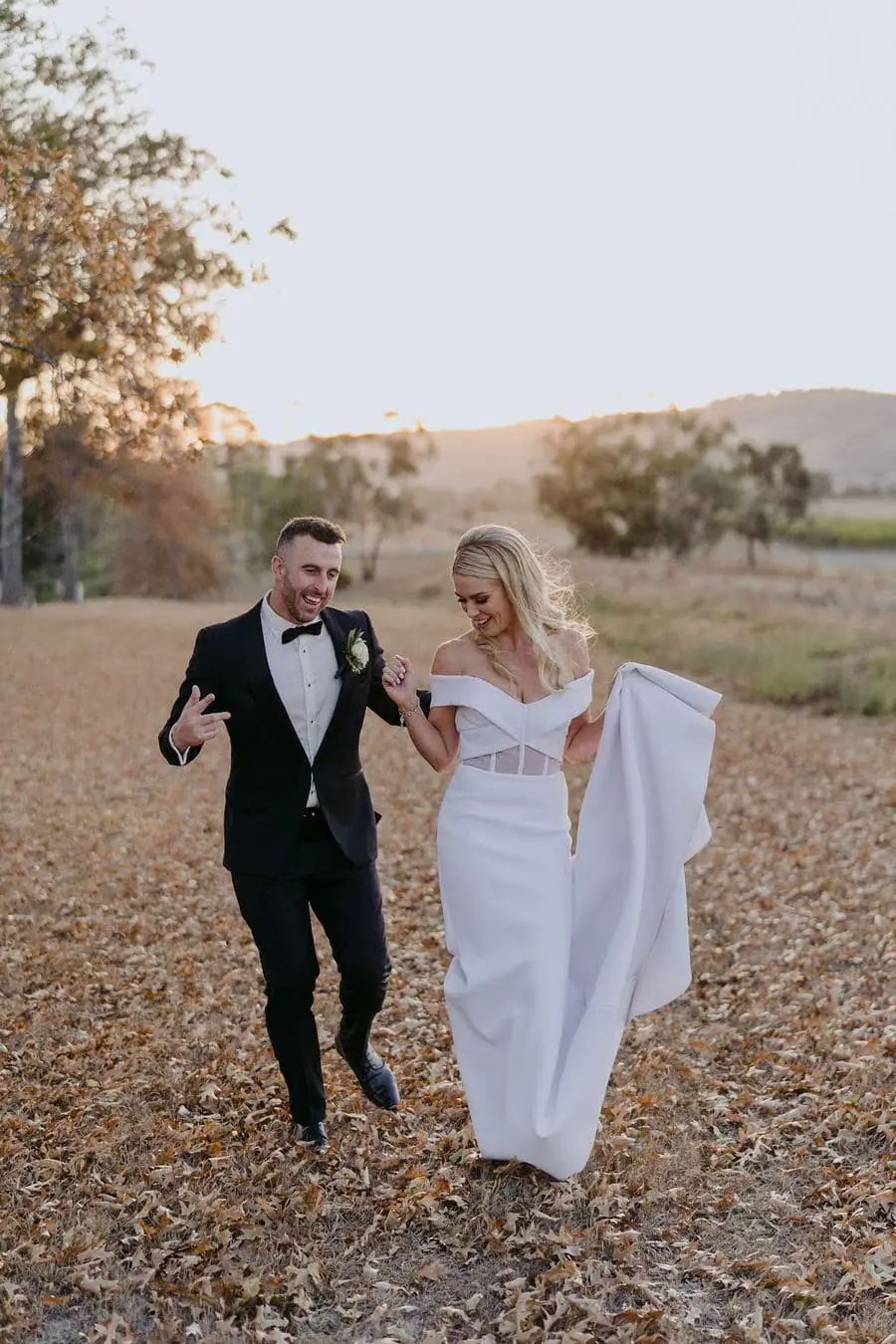 Couple walks through a dry field with the bride gathering her dress.