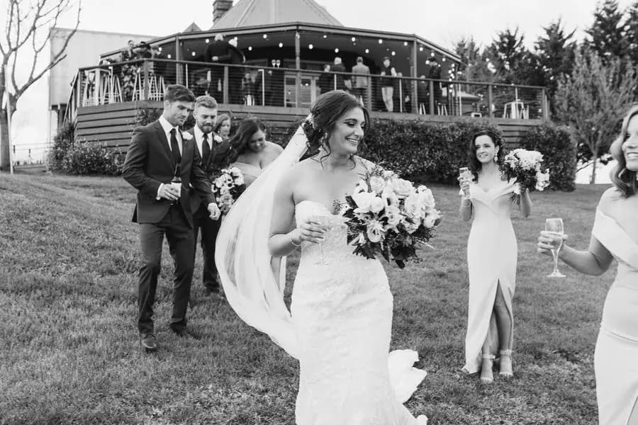Black and white photograph of the bride laughing with guests as the wedding party approaches behind her.