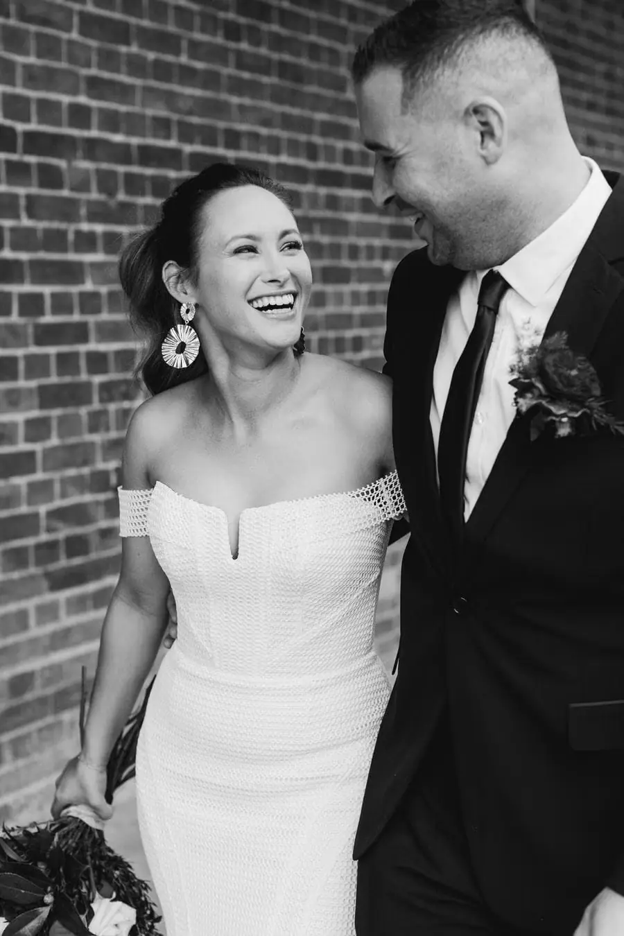 Black and white photograph of the couple laughing together beside a brick wall.