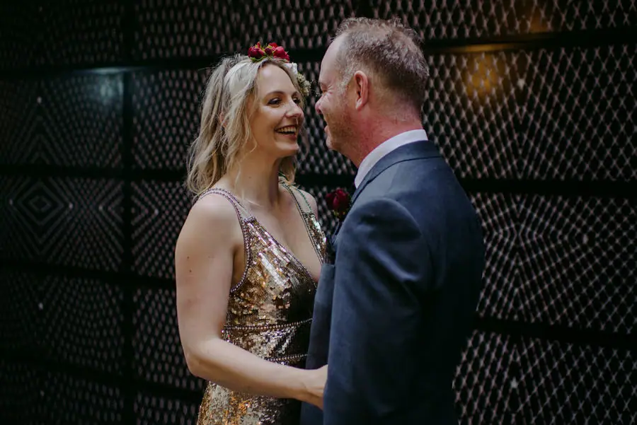 Couple faces each other smiling beside a patterned wall.