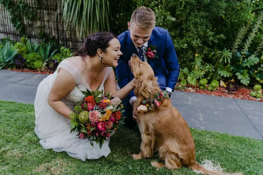 Couple crouches beside a dog wearing flowers in a garden.