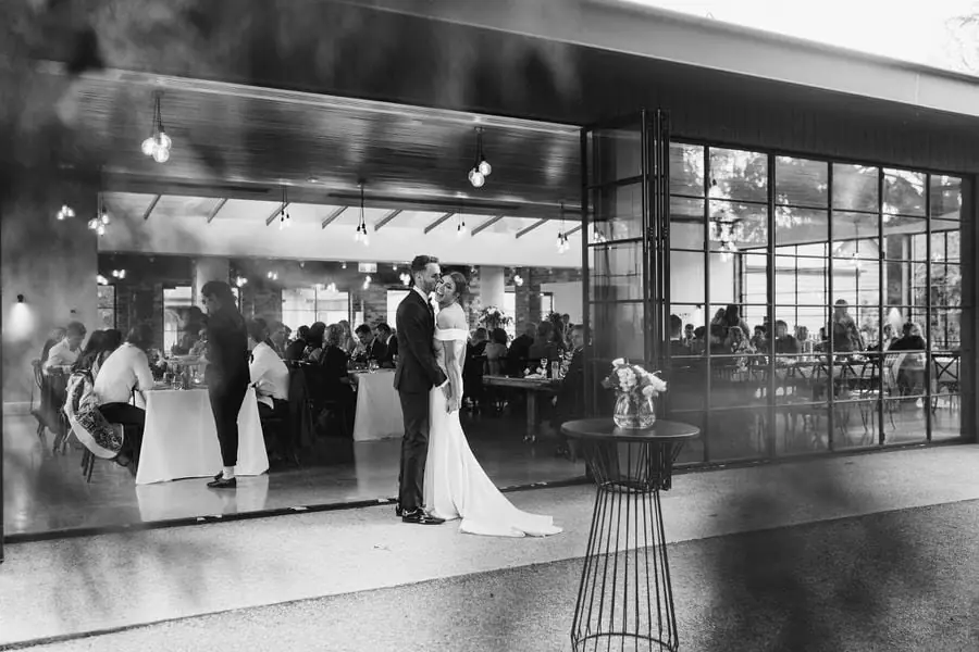 Black and white photograph of the couple embracing outside a glass-fronted reception venue.