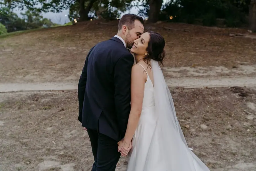 Couple kisses while walking hand in hand beside a wooded slope.