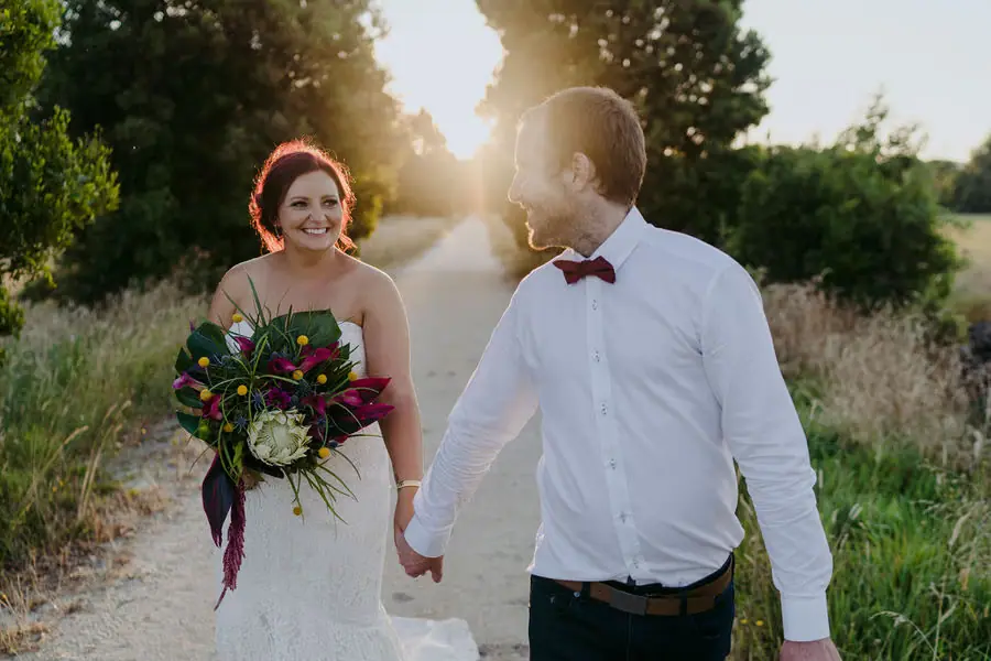Couple walks hand in hand along a sunny path with the bride holding a colourful bouquet.