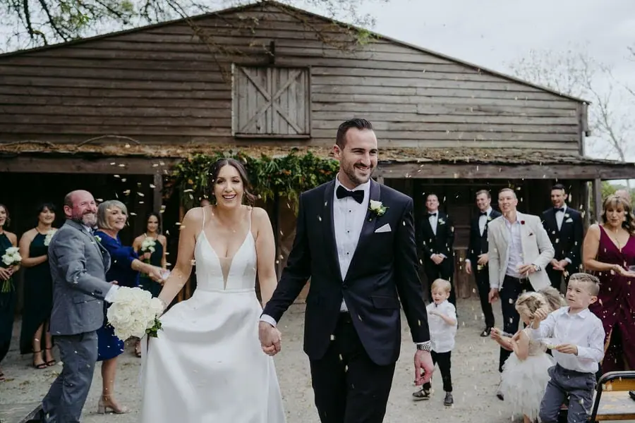 Newlyweds walk from a wooden venue through guests tossing confetti.