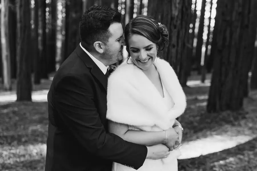 Black and white photograph of the groom embracing the bride from behind in a forest.