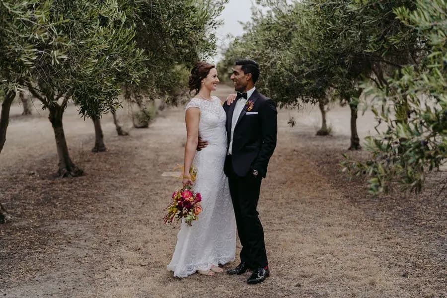 Couple smiles toward each other while standing between rows of trees.