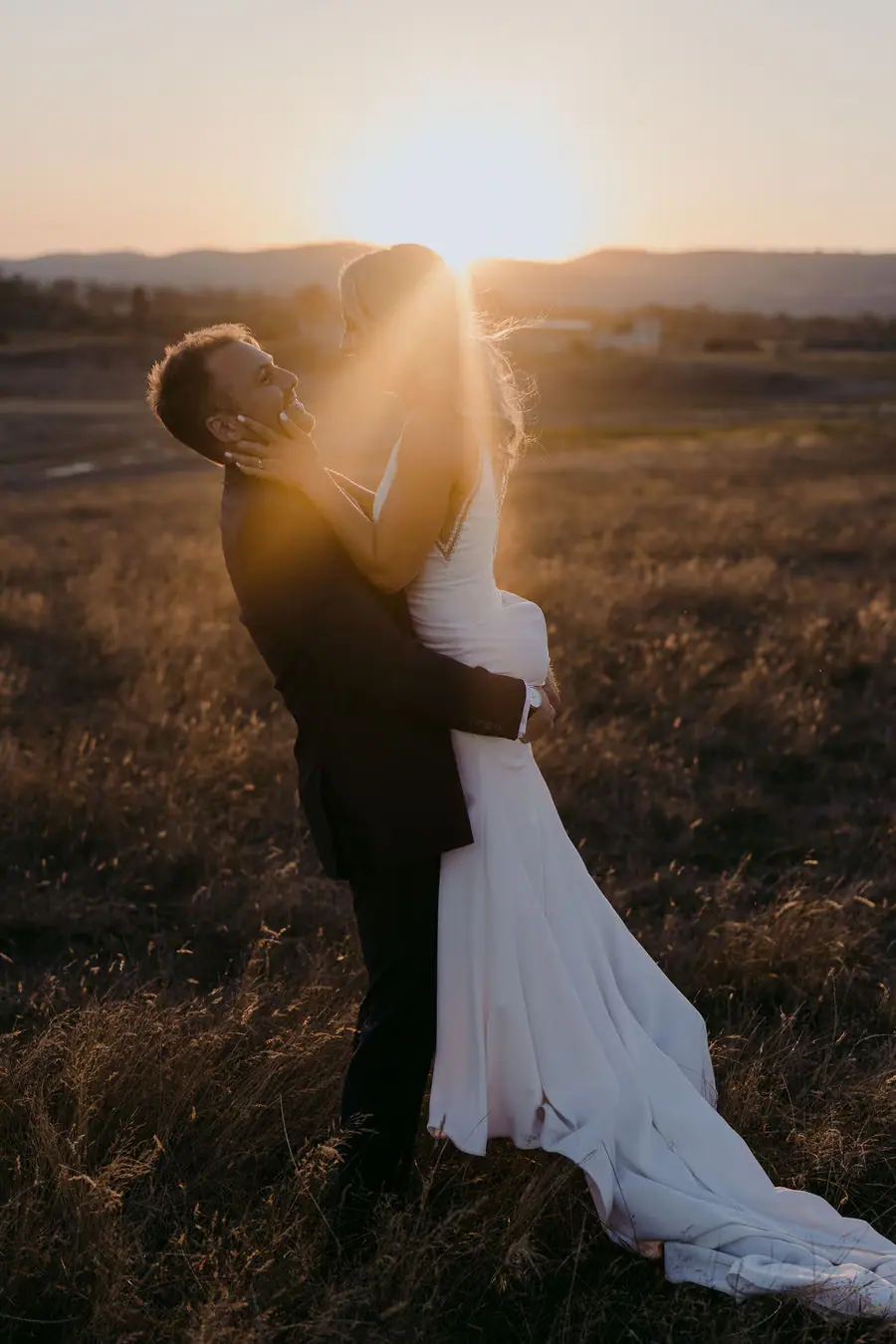 Couple embraces in a field with sunset light shining through the bride's raised arms.