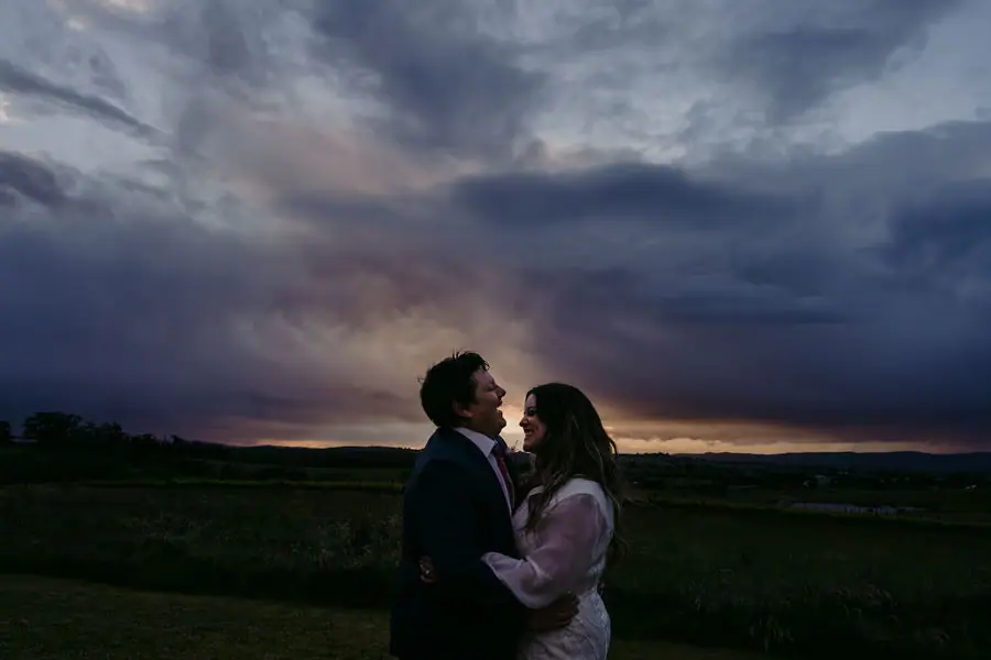 Couple embraces beneath a dramatic cloudy evening sky.