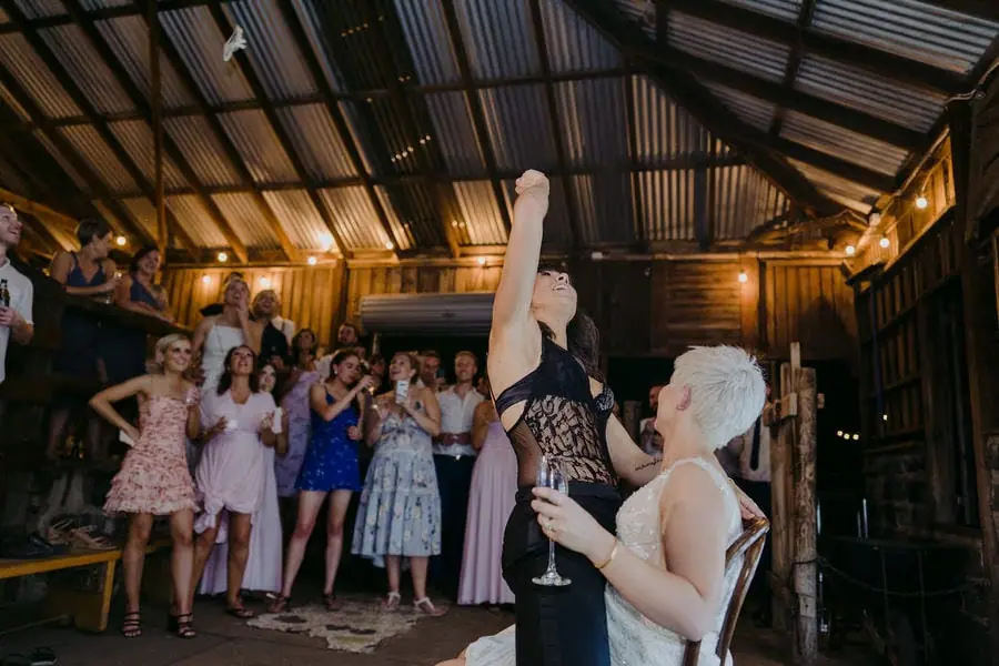Guests celebrate around a woman raising an arm inside a wooden reception venue.