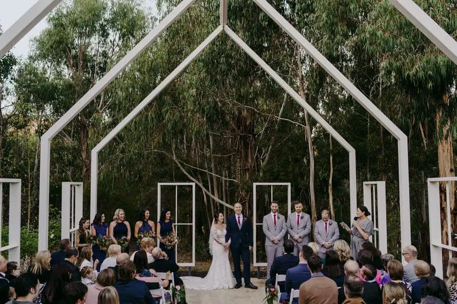 Wedding ceremony beneath open white roof beams surrounded by trees.