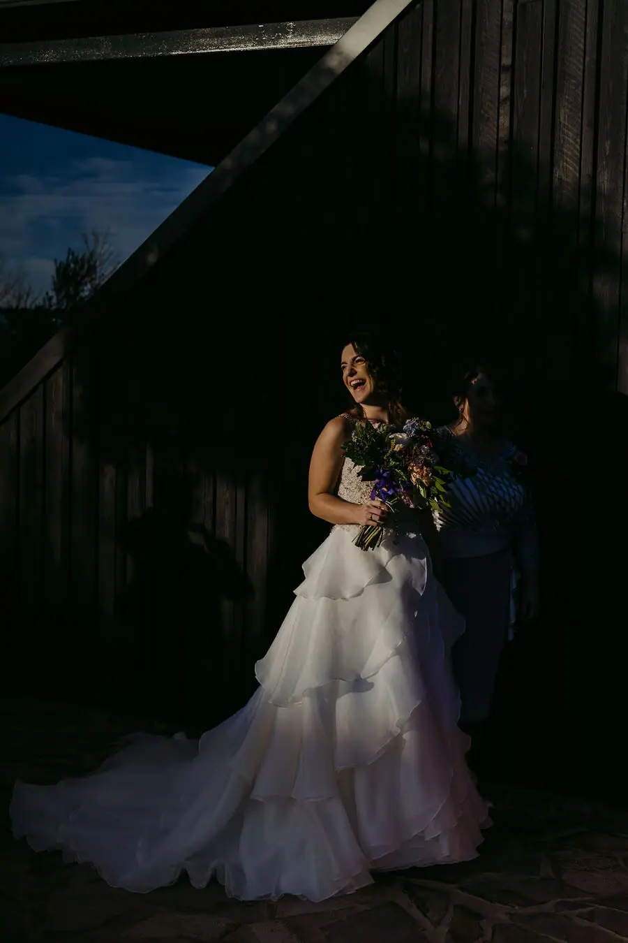 Bride holds a colourful bouquet in a pool of light beside a dark building.